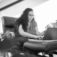 Working from home, a woman in her mid-thirties with a mixed racial background types on her laptop while seated on a chair, dressed comfortably.